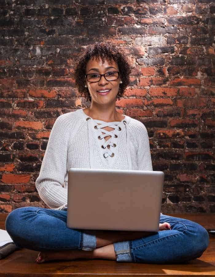 A cheerful woman wearing glasses and a white lace-up sweater sits cross-legged on the floor with a laptop on her lap. She’s working in front of a rustic brick wall, giving off a cozy and creative freelance writer vibe.