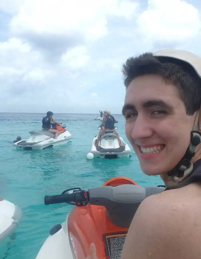 Smiling young man on a jet ski in turquoise ocean water, with other riders on jet skis in the background under a cloudy sky.