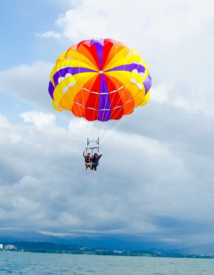 Two people parasailing high above the ocean, harnessed beneath a bright red, orange, yellow, and purple parachute with a backdrop of dramatic clouds and distant coastline.