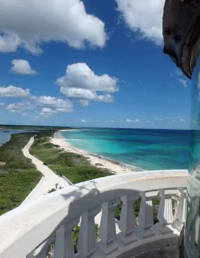 cenic view from the top of the Punta Sur lighthouse in Cozumel, showing a winding coastal road, lush greenery, and a pristine white-sand beach beside clear turquoise waters under a partly cloudy blue sky.