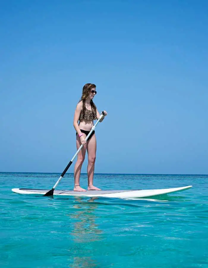 Woman in a bikini stand-up paddleboarding on calm, clear turquoise water under a cloudless blue sky, enjoying a peaceful day at sea.