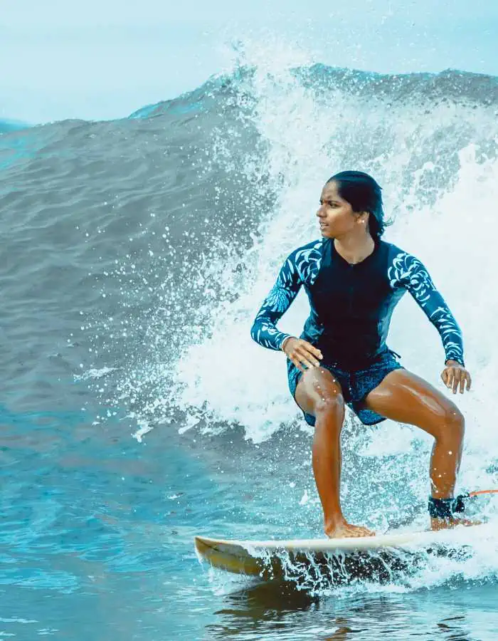 Surfer riding a wave with confident posture, wearing a blue and black rash guard and board shorts, water splashing behind them under overcast skies during a surfing lesson.