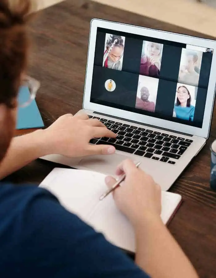 a person using a laptop to participate in a virtual meeting with five visible participants. The person is taking notes in a notebook while looking at the screen, suggesting they are actively engaged in the online discussion. A cup and a folder can be seen on the desk, indicating a workspace setup.