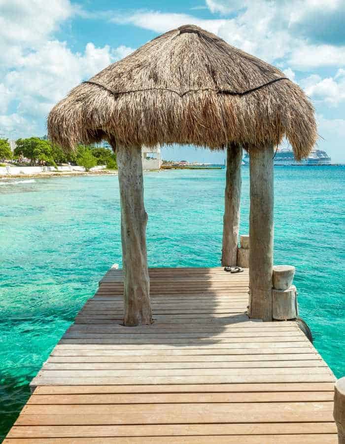 A wooden pier extending over clear turquoise water, ending in a small palapa (a thatched-roof shelter). The serene, tropical setting includes a glimpse of the shoreline in the background, under a partly cloudy sky.