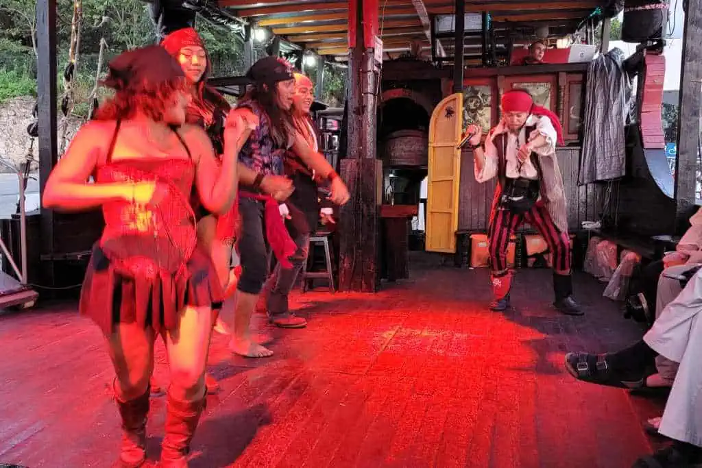 Performers dressed as pirates entertain guests with a lively dance aboard a themed ship. The stage is bathed in red lighting, enhancing the energetic atmosphere as the dancers, wearing pirate-inspired costumes with bandanas and boots, move in sync. A pirate captain, dressed in a long coat and hat, engages the audience with a theatrical pose.