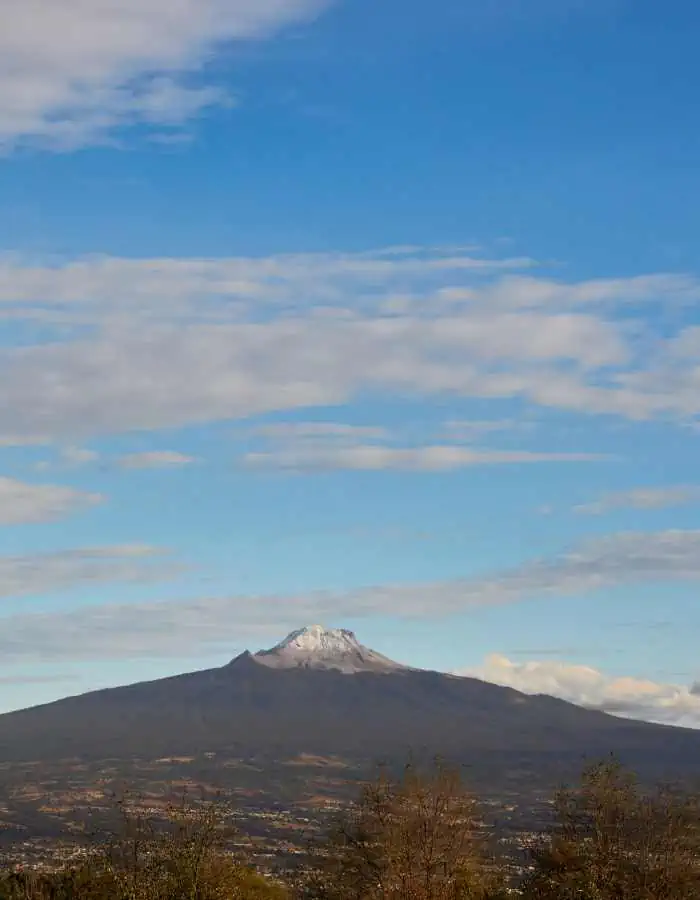 Scenic daytime view of La Malinche volcano, with its partially snow-dusted summit rising above the surrounding landscape under a sky dotted with white clouds.