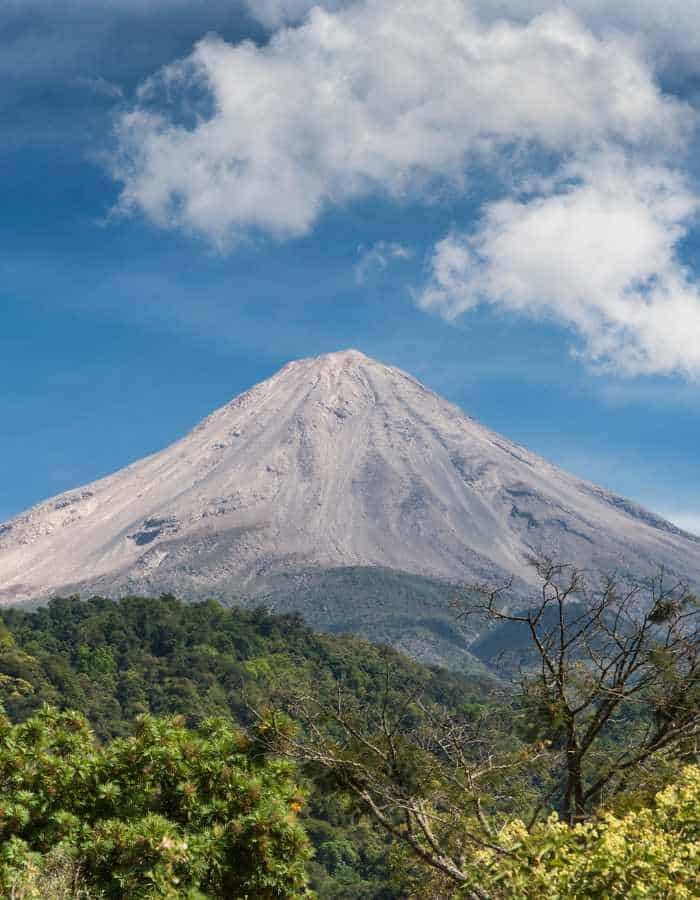 Volcán de Colima, also known as Volcán de Fuego, stands tall with its ash-covered slopes against a backdrop of fluffy clouds and a vibrant blue sky, framed by lush green foliage in the foreground.