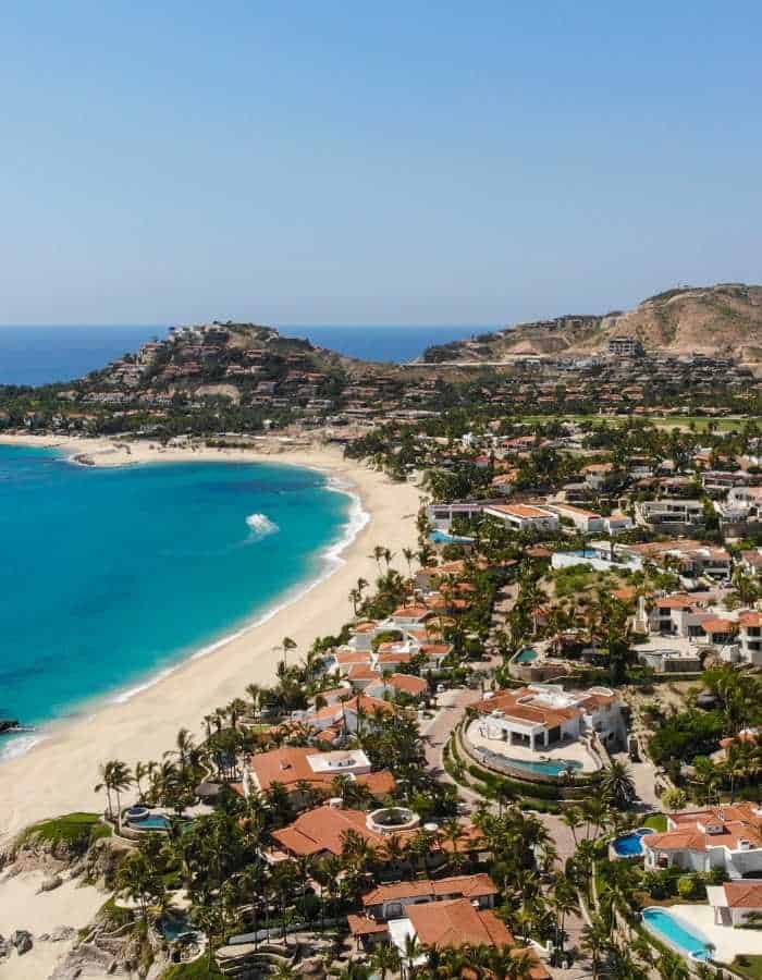 Aerial shot of a picturesque Cabo beachfront neighborhood with red-roofed houses, palm trees, a clear sandy beach, and a boat cruising near the shore.