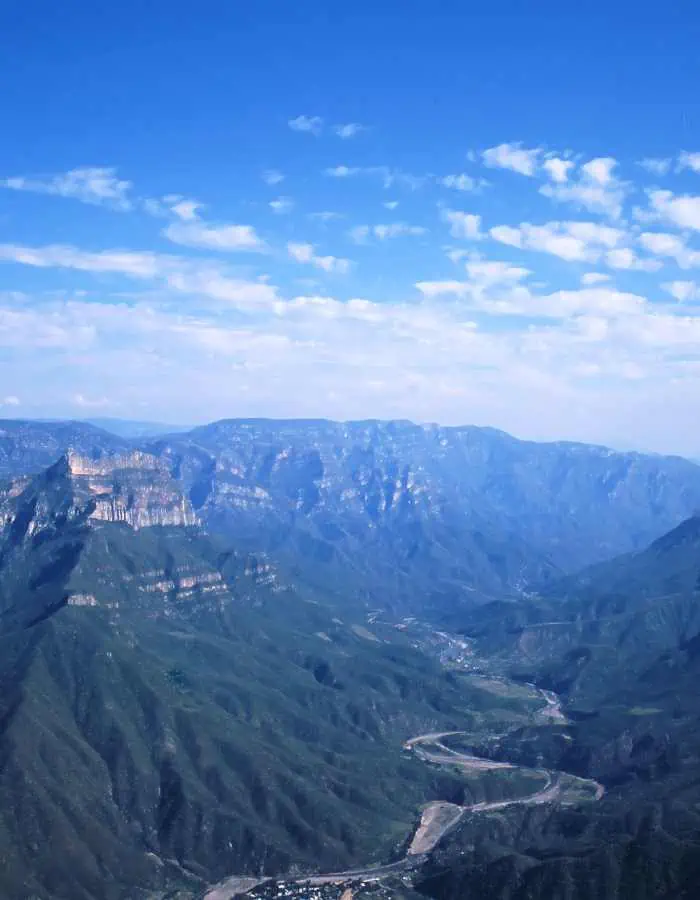 Breathtaking aerial view of Copper Canyon in Mexico, with deep valleys and sheer cliff faces, verdant with greenery, under a vast blue sky with wispy clouds.