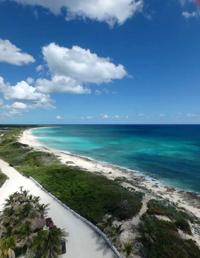 Aerial view of the serene Punta Sur coastline in Cozumel, showcasing the white sandy beach, lush greenery, and clear turquoise waters under a bright blue sky with scattered clouds.
