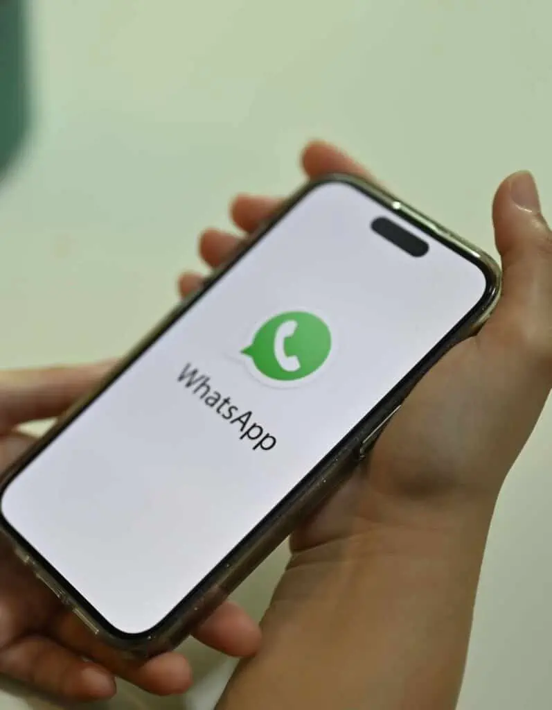 A person holds a smartphone displaying the WhatsApp logo on the screen. The image focuses on the phone's screen, showing the app's white background with a green speech bubble and phone icon, indicating the app is loading or opening.