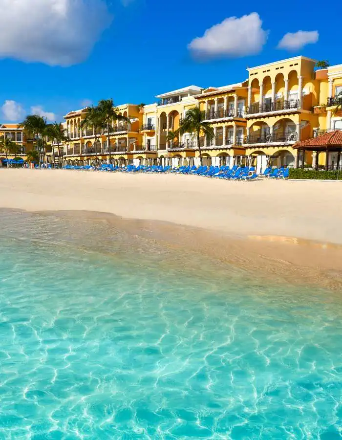 The idyllic Playa del Carmen beachfront, featuring rows of blue loungers and parasols on powdery white sands, with the crystal-clear Caribbean waters in the foreground and a luxurious hotel boasting Spanish-style architecture in the background.