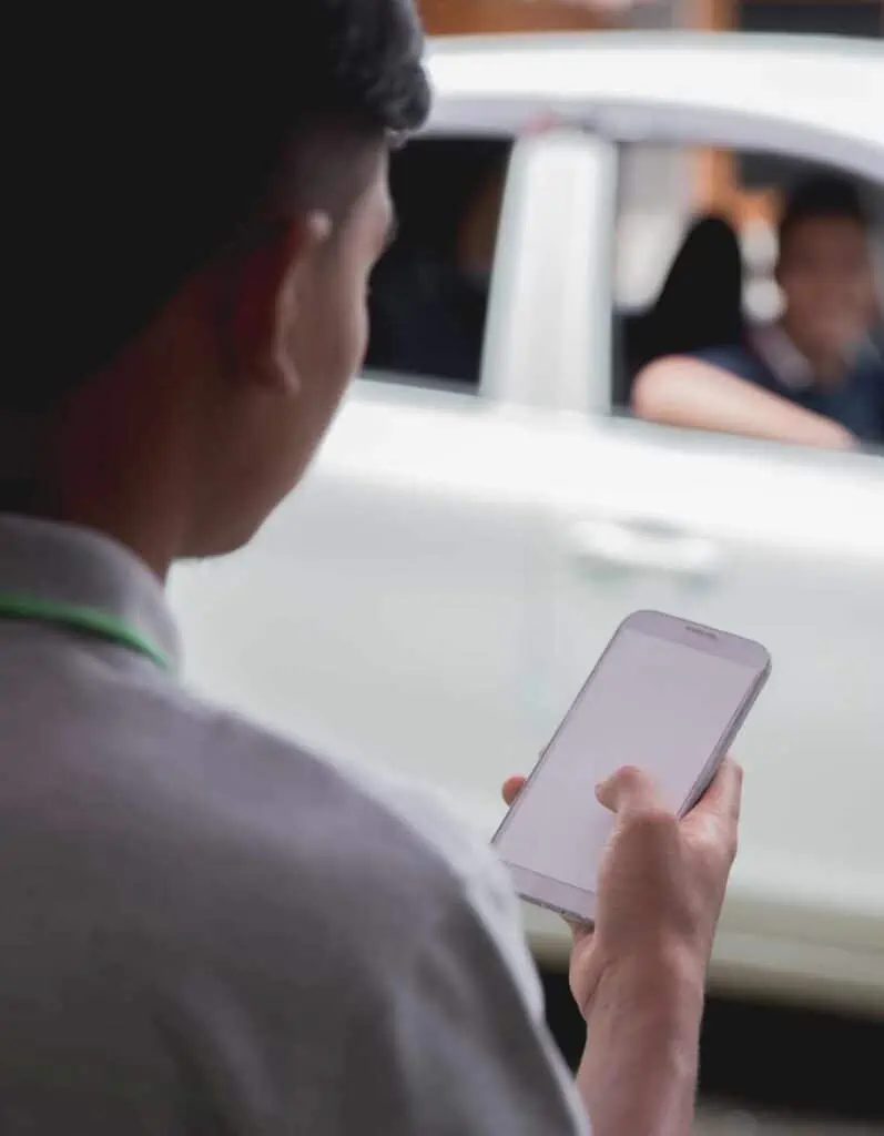 Over-the-shoulder view of a man holding a smartphone with a blank screen, with the background showing a blurred image of a taxi and its passengers, depicting a common scene of urban connectivity and ride-hailing.