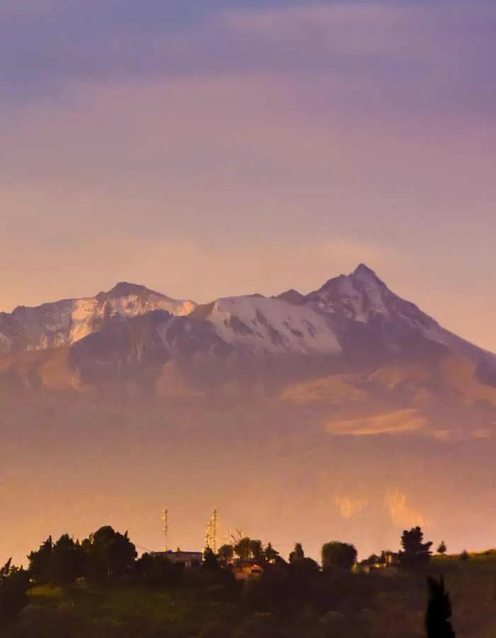 Early morning view of Nevado de Toluca with sunlight casting a warm glow on its snow-capped peak, silhouette of communication towers and tree line in the foreground.