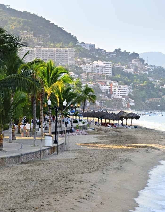 The beachfront of Puerto Vallarta at dusk, with lush palm trees lining the promenade, thatched beach umbrellas, and a gentle surf, all set against a backdrop of the city's hillside buildings gradually fading into the misty tropical hills.