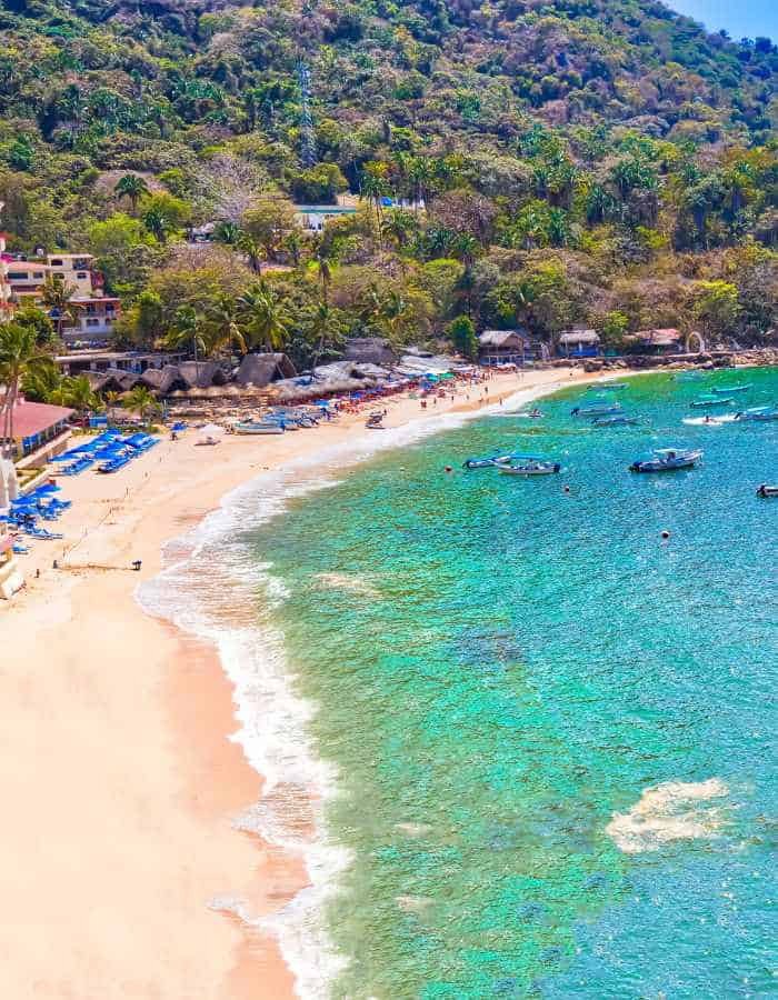 A picturesque curve of sandy beach in Puerto Vallarta, with beachgoers and blue parasols dotted along the shore, small boats floating in the clear turquoise water, all nestled against a lush, green mountain backdrop, embodying a vibrant coastal getaway.