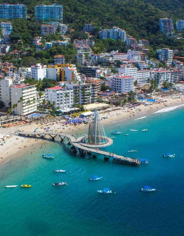 An aerial shot captures the bustling Los Muertos Beach in Puerto Vallarta, featuring the iconic pier with its sail-like structure, surrounded by azure waters, colorful beach umbrellas, and nestled at the foot of the verdant Sierra Madre mountains.