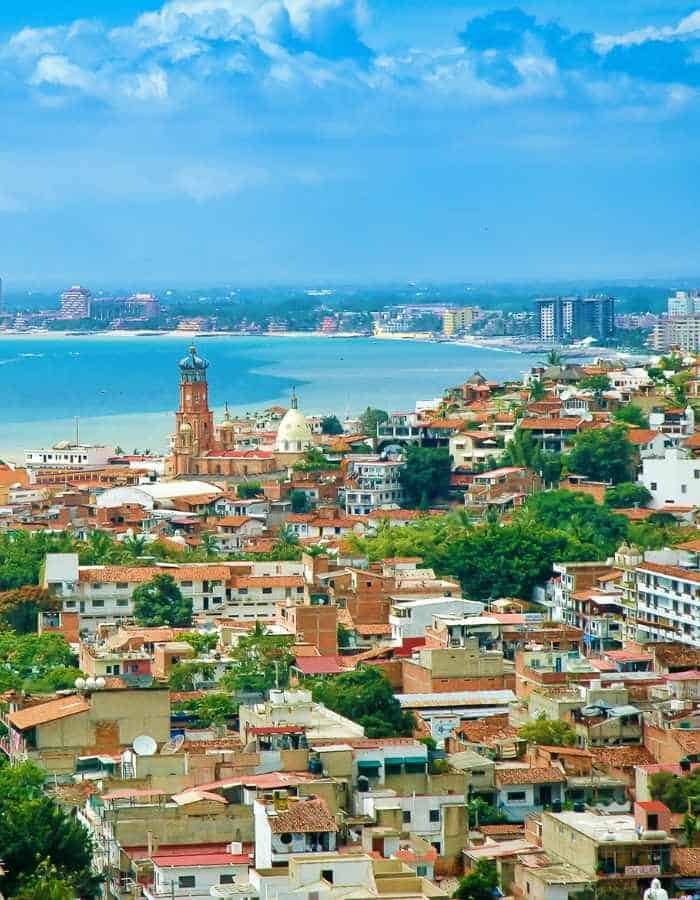 A vibrant view over the terracotta rooftops of Puerto Vallarta, with the iconic Church of Our Lady of Guadalupe tower standing out against a backdrop of lush green hills, overlooking the calm blue waters of Banderas Bay.