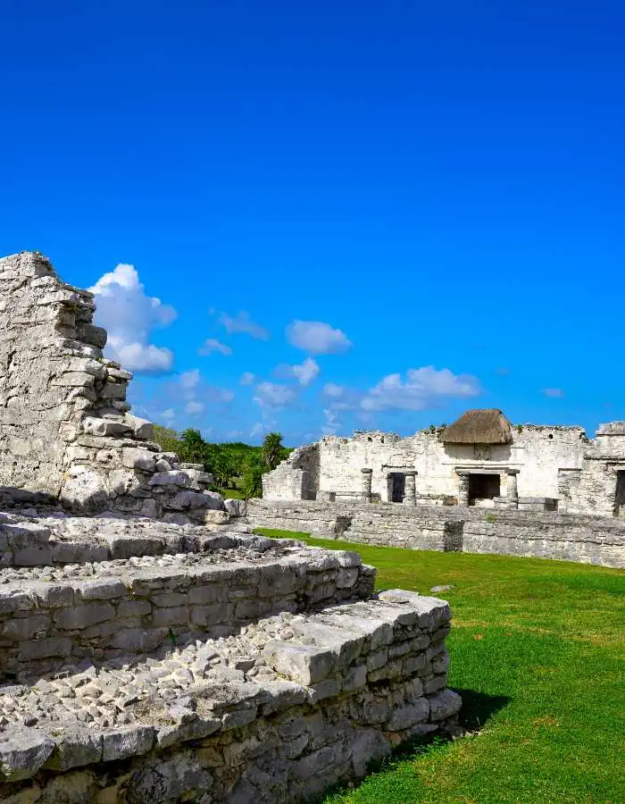 Ancient Mayan ruins of Tulum set against a vivid blue sky, showcasing the contrast between the well-preserved stone structures with a thatched roof in the background and the lush green grass in the foreground, a testament to the rich history of the Yucatan Peninsula.