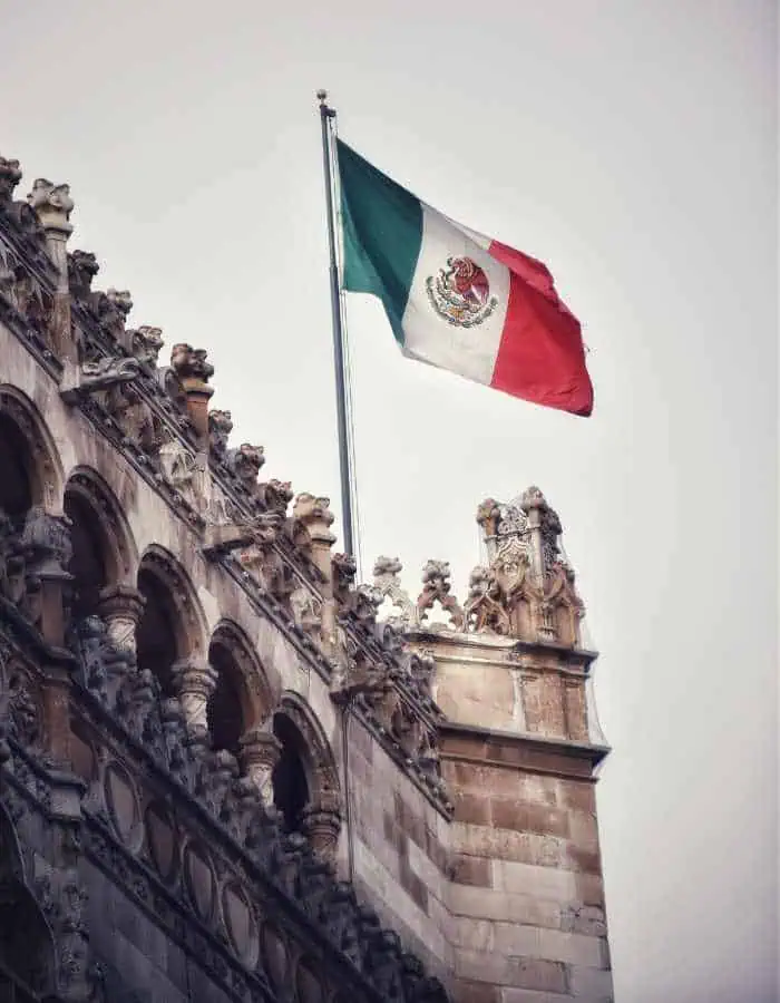 A Mexican flag hoisted on a pole, billowing against a cloudy sky, contrasts with the intricate Gothic-style architecture of a stone building adorned with numerous statues. The juxtaposition of national symbolism with historical architecture conveys a sense of heritage and pride.