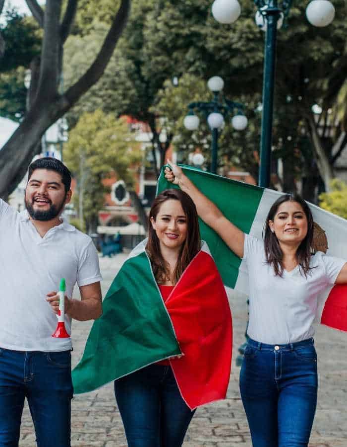 Three cheerful individuals are celebrating, each wearing white shirts and jeans, with the Mexican flag proudly displayed. The person in the center is draped in the flag, while the others wave their flags in the air, evoking a sense of national pride and joy.