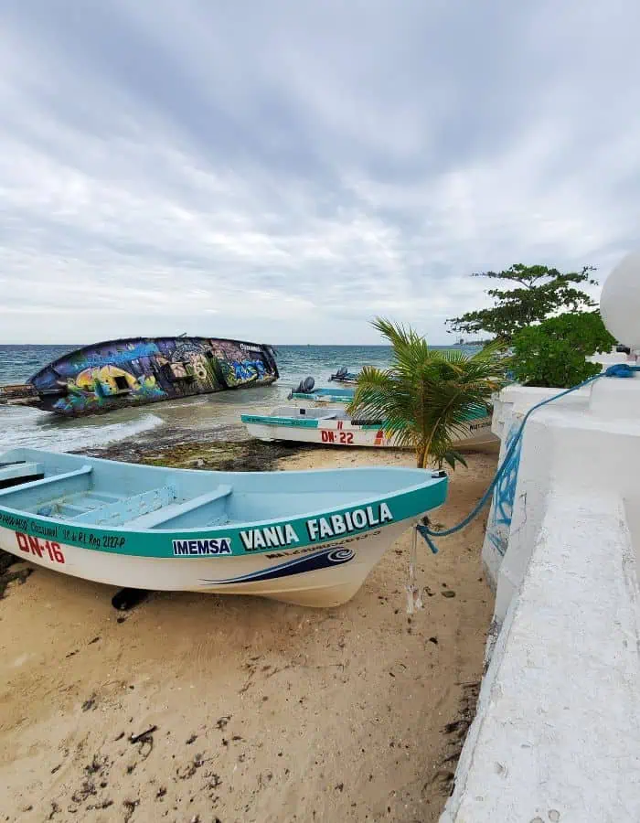 Small fishing boats rest on the sandy shore of Cozumel, including one named "Vania Fabiola," with a palm tree and white wall nearby. In the background, a large, overturned boat covered in colorful graffiti lies partially in the surf under a cloudy sky.