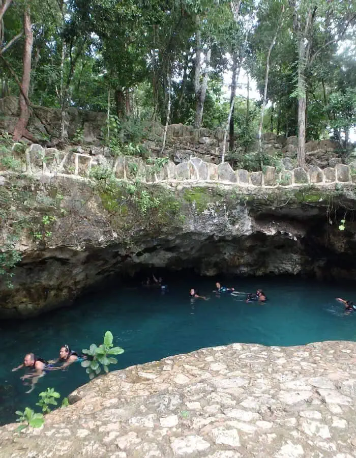 This image showcases a breathtaking open-air cenote at Cenotes Casa Tortuga in Tulum, Mexico. The turquoise water is surrounded by rugged limestone formations, with swimmers wearing life jackets exploring the refreshing natural pool. Lush greenery and trees grow along the rocky edges, blending seamlessly with the jungle landscape.