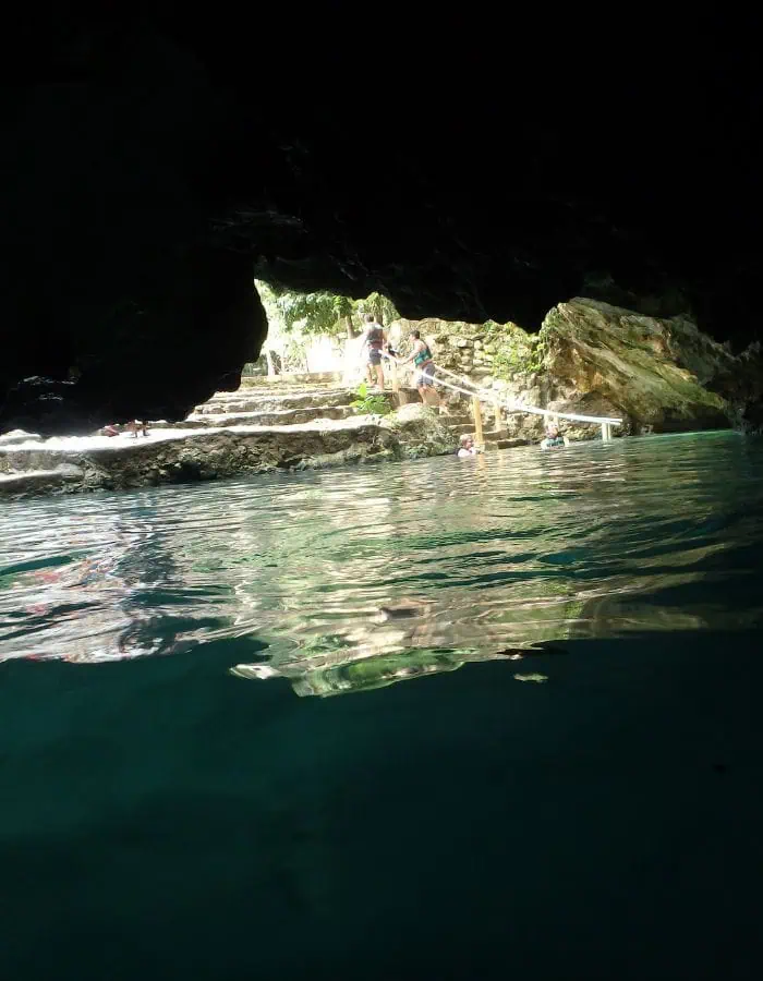 This image captures the stunning view from inside a cenote cave at Cenotes Casa Tortuga in Tulum, Mexico. The dark rocky entrance frames the bright outdoor area where visitors are seen stepping into the water from stone stairs.