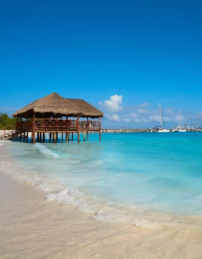 A peaceful beach scene at Playa Maroma in Mexico, featuring a thatched-roof wooden pavilion built over the water. The turquoise sea gently laps against the white sand shoreline, while sailboats and a marina are visible in the distance under a vibrant blue sky.