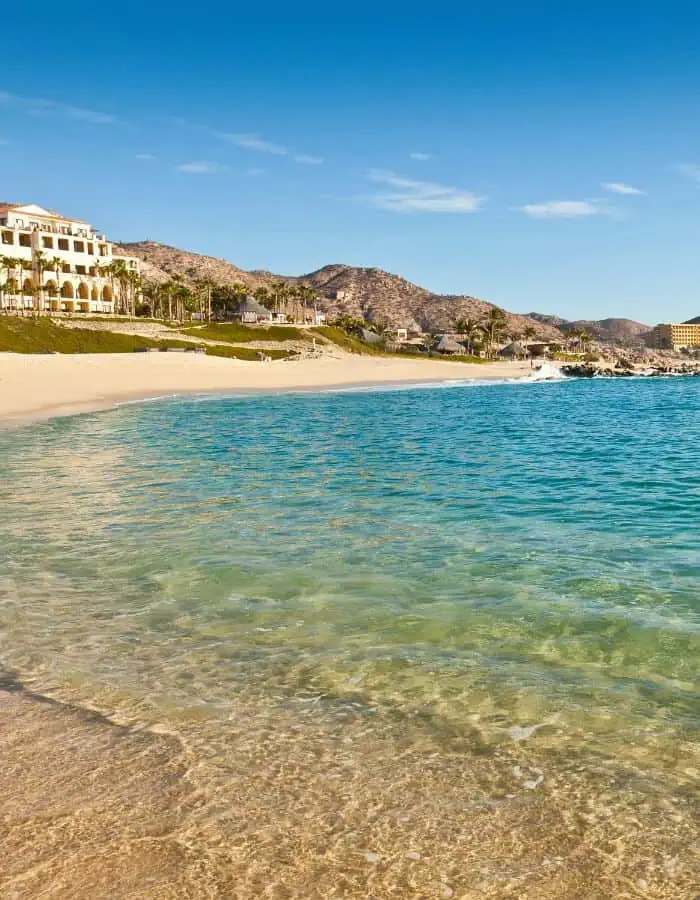 A serene beach in Cabo San Lucas, Mexico, with clear, shallow turquoise water meeting soft golden sand. Elegant beachfront resorts line the shore, backed by arid, rugged hills under a bright blue sky.
