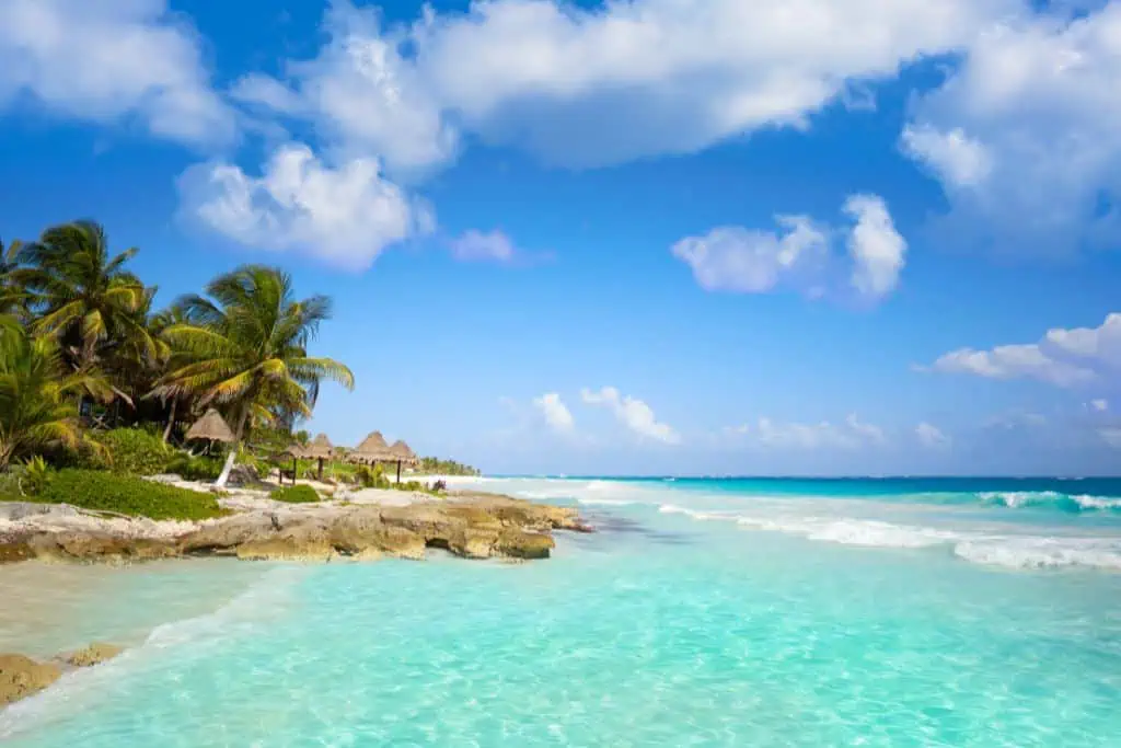 Turquoise waves wash up against the rocky shoreline of Paamul Beach, with palm trees and thatched-roof palapas nestled among the greenery. A bright blue sky with scattered clouds completes the tropical, laid-back vibe of this coastal hideaway.