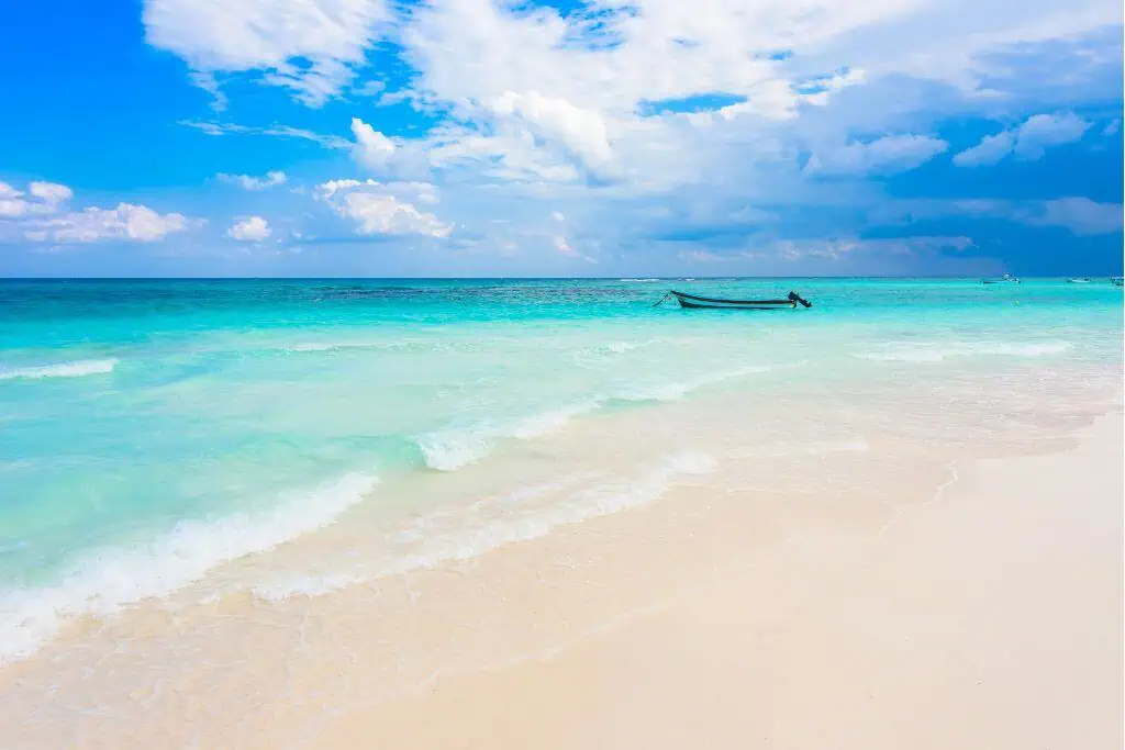 A tranquil beach scene with soft white sand in the foreground and turquoise waves gently washing ashore under a vibrant blue sky. A small boat floats near the horizon, adding a peaceful touch to the tropical seascape.