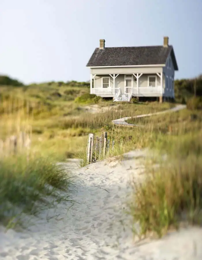 Cozy beach house with a wide front porch, white railings, and wooden steps leading up from a winding sandy path through grassy dunes. The sky is clear, and the house sits peacefully in a sunlit coastal landscape.