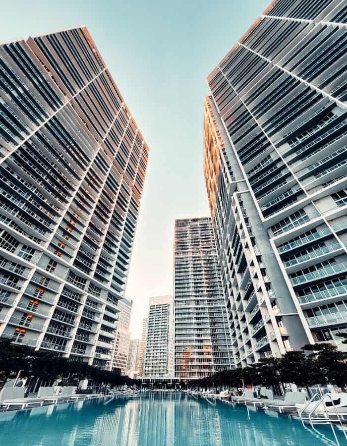 Tall, modern high-rise condominiums with rows of balconies frame a central outdoor pool, reflecting the glassy facades and early evening sky. Lounge chairs line the poolside, while the symmetrical architecture and crisp lines create a luxe, urban resort vibe.
