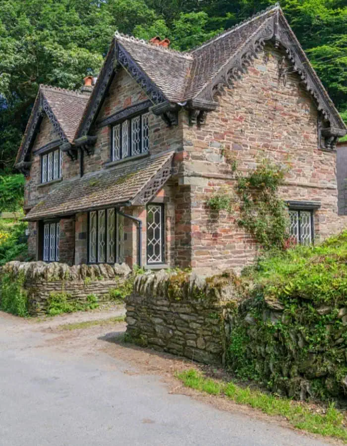 Charming stone cottage with steep gabled roofs, decorative trim, and tall diamond-paned windows, nestled by a narrow country road. Ivy climbs the rustic stone walls, and the house is framed by lush greenery and a low mossy stone fence.
