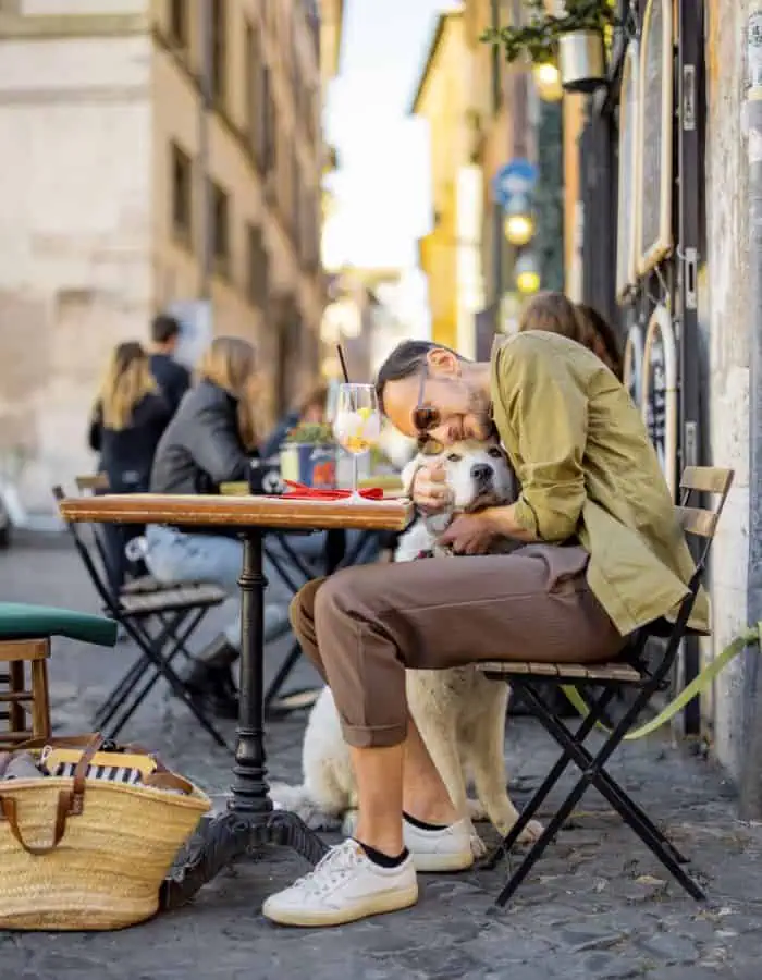 A man in sunglasses and casual clothes sits at a bistro table on a cobblestone street, lovingly hugging a large white dog beside him while sipping an orange cocktail. The cozy café scene is full of warmth and charm, with other diners in the background and a woven straw bag resting at the base of the table.