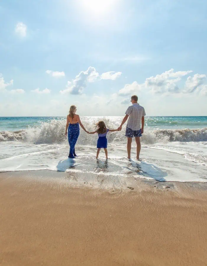 A family of three—two adults and a young girl—stand hand-in-hand at the edge of a beach, facing the ocean as waves crash toward them under a bright, sunny sky.