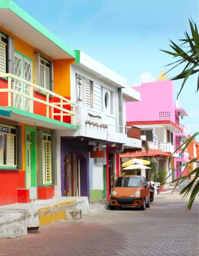 Colorful street scene in downtown Isla Mujeres featuring vibrant buildings painted in red, green, orange, purple, and pink hues, with a small orange car parked along a narrow cobblestone road under a bright blue sky.