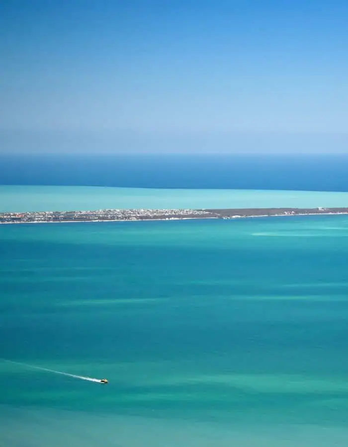 An aerial view of a long, narrow island surrounded by striking shades of turquoise and deep blue ocean waters, with a small yellow boat leaving a white trail across the sea in the foreground under a clear blue sky.