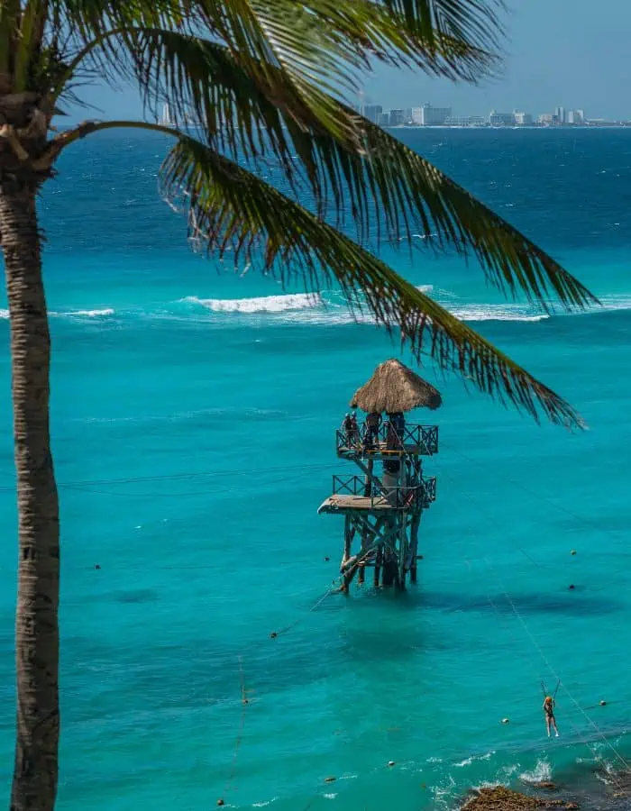 A scenic view of a tall wooden tower with a thatched roof standing in the middle of vibrant turquoise waters, likely part of a zipline attraction. A person is seen ziplining toward the shore, while a palm tree frames the foreground and a distant city skyline stretches across the background.