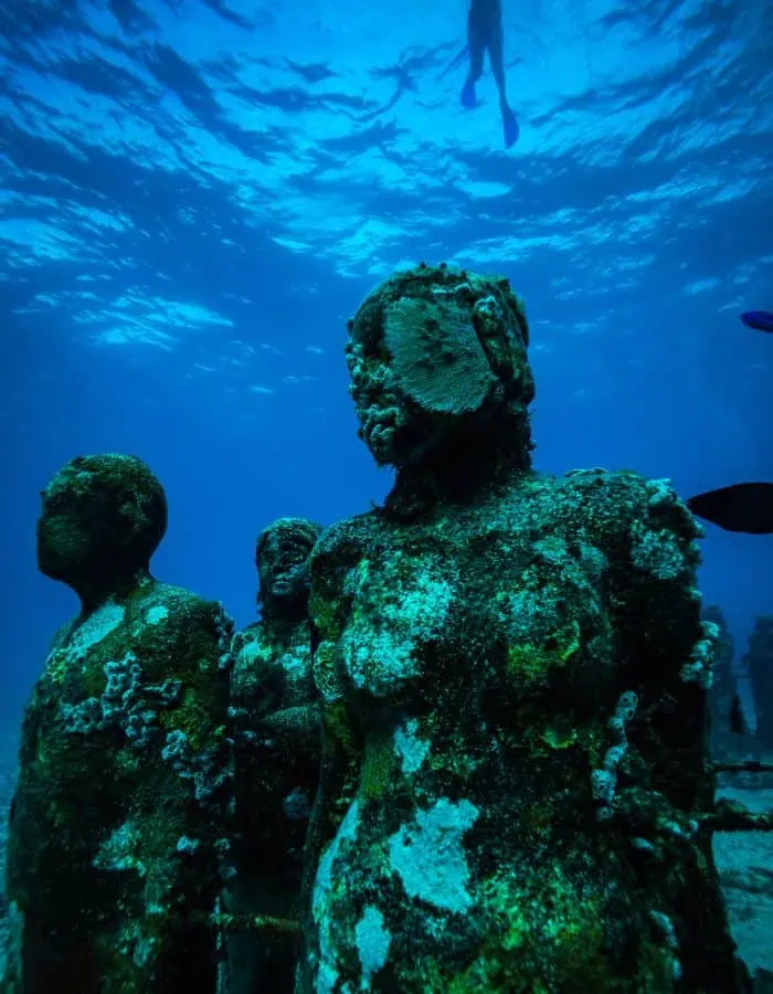 Underwater view of algae-covered human statues standing on the ocean floor at the MUSA underwater museum, with a snorkeler and fish swimming above in the clear blue water.