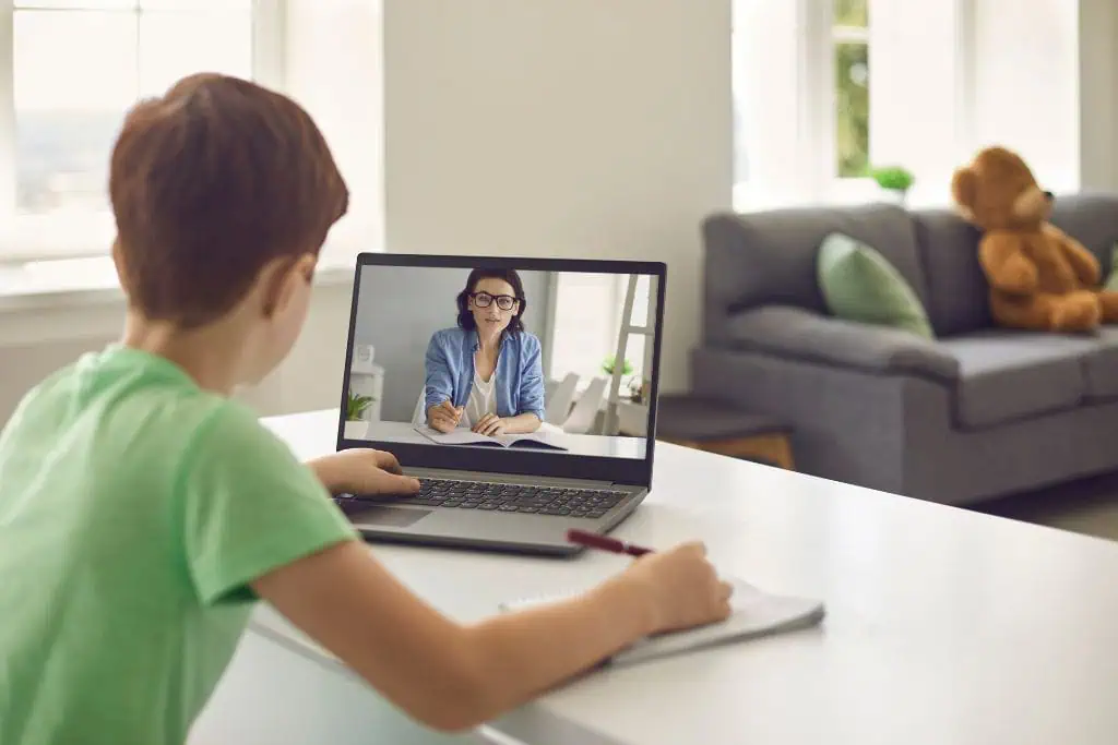 A young boy in a green shirt participates in an online class, attentively watching a female teacher on his laptop screen while taking notes in a notebook. The scene is set in a bright, cozy living room with a grey couch and a large teddy bear in the background.