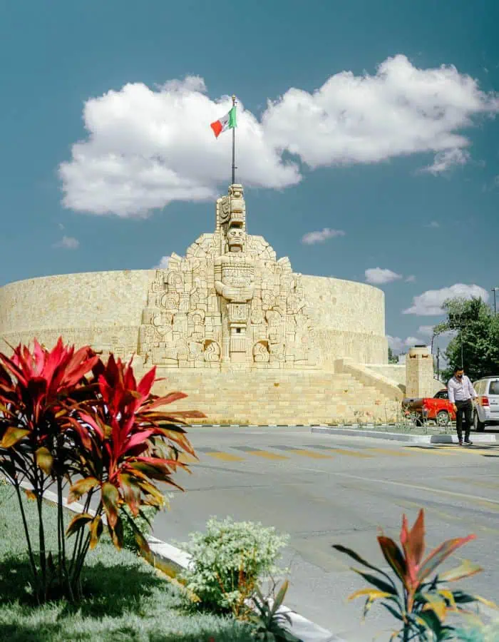A wide-angle shot of the Monumento a la Patria in Mérida, Mexico, a large stone sculpture featuring intricate carvings of Mayan figures and symbols, with a Mexican flag flying above. The monument sits in a circular stone base surrounded by a landscaped street, with red tropical plants in the foreground and a man crossing the road to the right under a blue sky with puffy clouds.