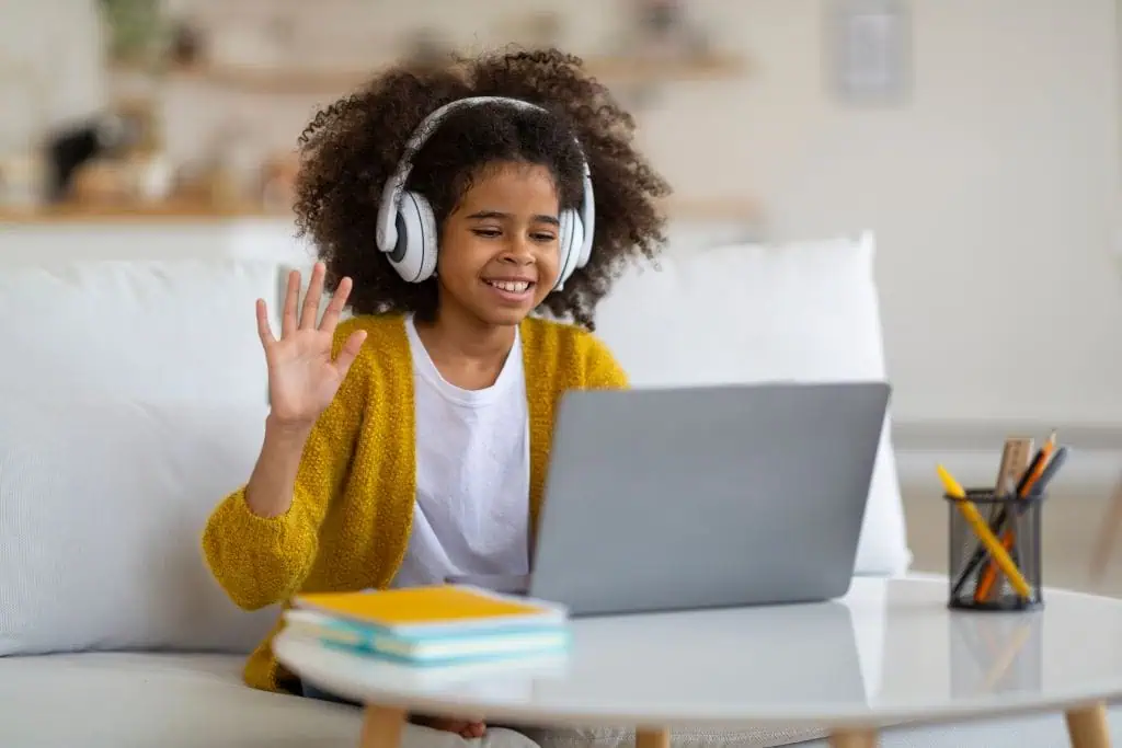 Smiling girl wearing headphones and a mustard yellow cardigan waves at her laptop during an online class while sitting on a white couch. A few notebooks and a pencil holder sit on the coffee table in front of her, creating a cozy and cheerful virtual learning setup.