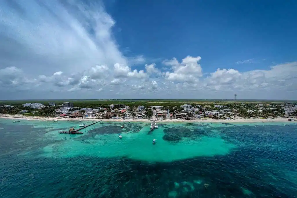 Aerial view of Puerto Morelos showcasing a charming beachfront town with a central pier extending into vibrant turquoise waters. Coral reefs are visible beneath the surface, surrounded by small boats and framed by a vast blue sky with scattered clouds.