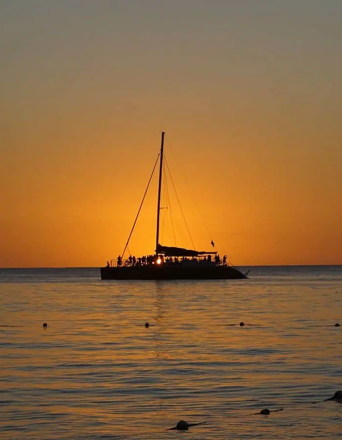 A silhouette of a catamaran filled with people sailing at sunset, with the orange and golden hues of the sky reflecting on the calm ocean water, creating a serene and picturesque scene.