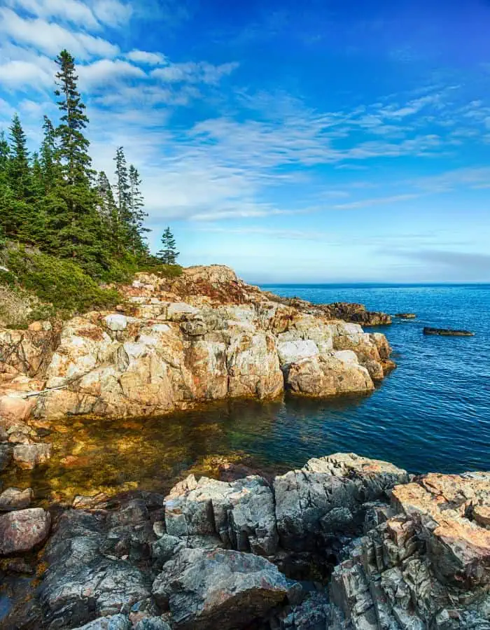 Jagged granite cliffs meet the deep blue Atlantic Ocean at Acadia National Park, with evergreen trees perched atop the rocky shoreline under a sky streaked with wispy clouds. The rugged coast and vibrant colors showcase the wild, scenic beauty of Maine’s coastal wilderness.