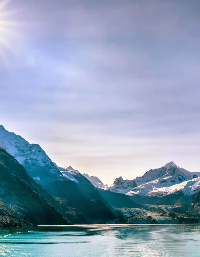 now-covered mountains rise above a turquoise glacial bay in Alaska, with sunlight casting a soft glow across the icy peaks and calm water. The dramatic contrast between the dark mountain shadows and the shimmering glacier highlights the untouched beauty of the Alaskan wilderness.