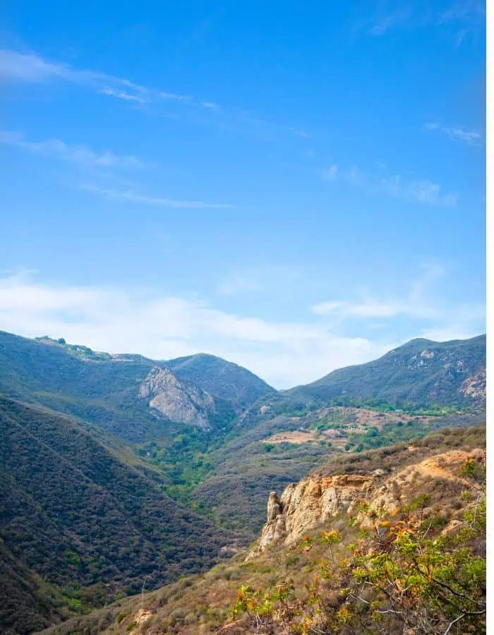 A scenic view of the Santa Monica Mountains under a clear blue sky, with rolling ridges, rocky outcrops, and patches of green vegetation scattered across the arid hills. The valley between the peaks creates a natural corridor, showcasing Southern California’s rugged coastal landscape.