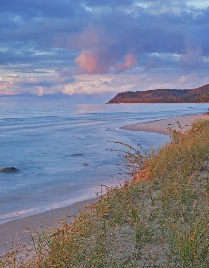 Grassy sand dunes gently slope toward the calm shoreline of Lake Michigan at Sleeping Bear Dunes National Lakeshore, with soft pastel clouds reflecting the warm glow of sunset. A distant forested bluff curves into the lake, adding depth and serenity to the peaceful coastal scene.